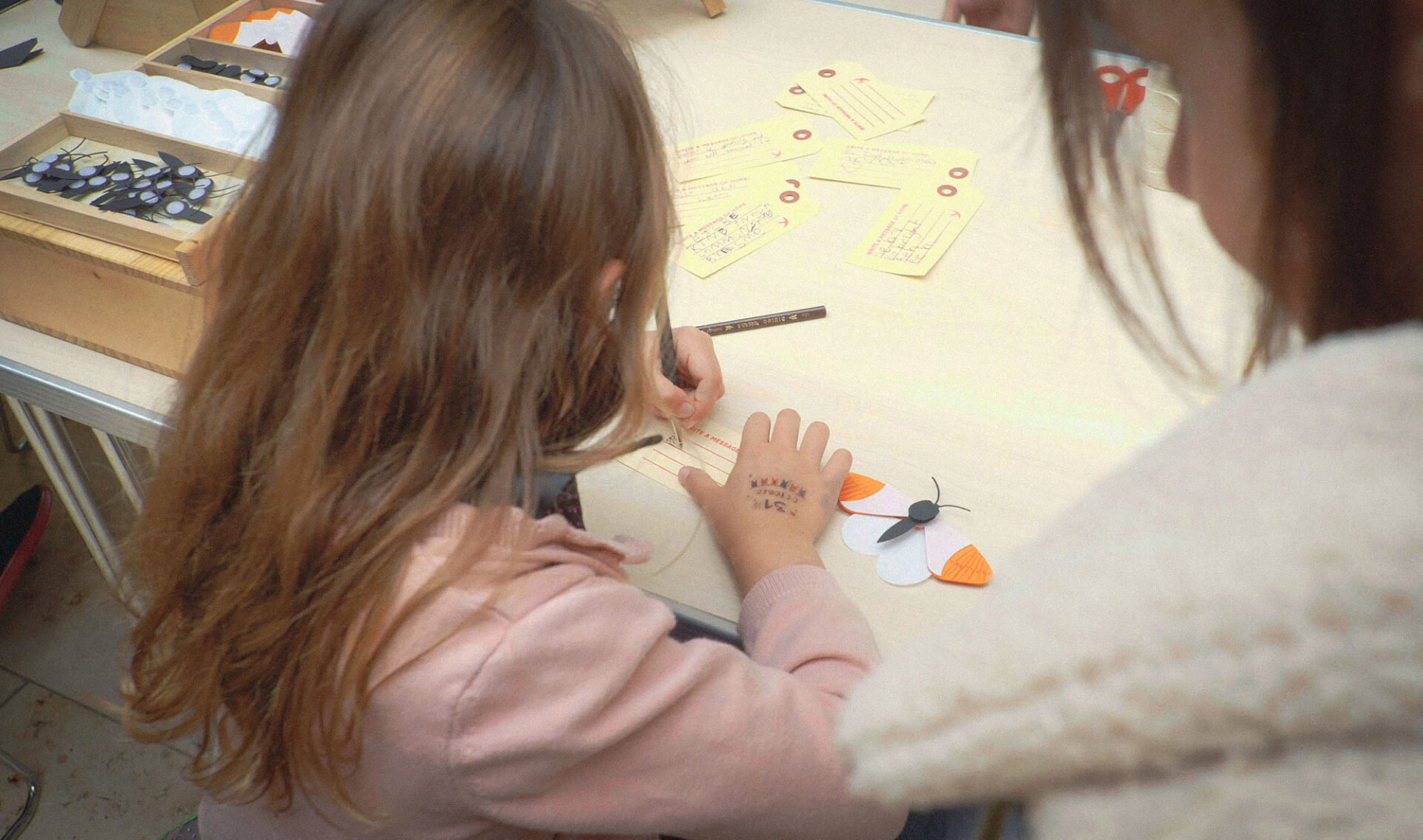 A young girl writes a message of hope on a luggage label with her mother watching over her.